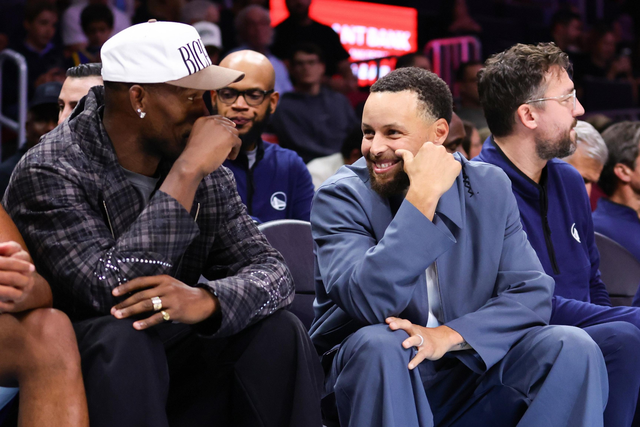 Two men seated courtside, one in a white cap and plaid jacket, the other in a blue suit, smiling and engaged in conversation.
